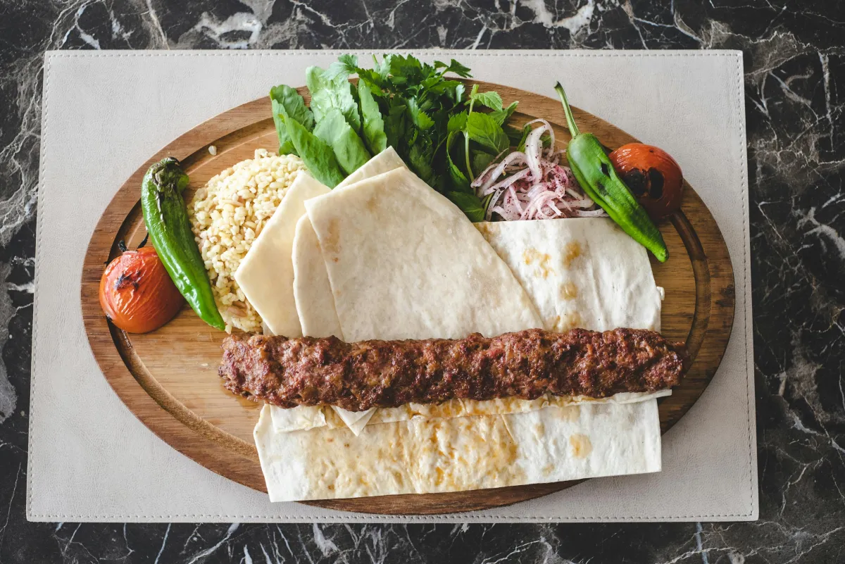 Adana kebab sur planche en bois avec lavaş, oignon rouge au sumac et tomates grillées
