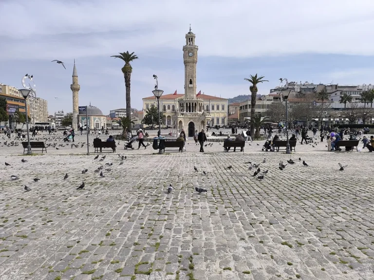Grande place pavée avec pigeons, palmiers et une tour de l'horloge ottomane au fond