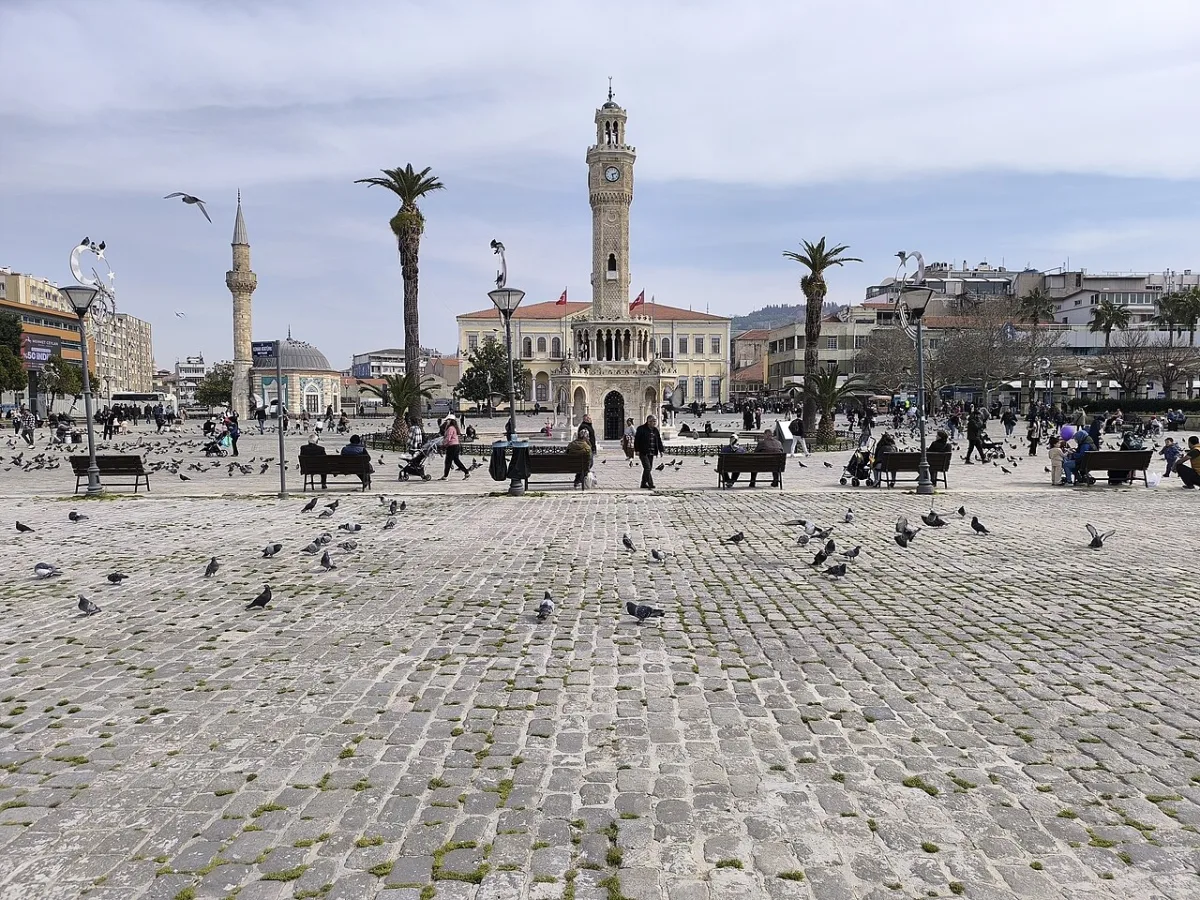 Grande place pavée avec pigeons, palmiers et une tour de l'horloge ottomane au fond