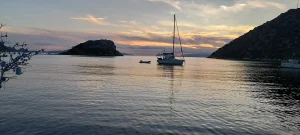 Coucher de soleil orange sur une baie calme avec un voilier au mouillage et un îlot rocheux qui ferme l'horizon