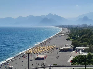 Longue plage rectiligne au revêtement gris bordée de parasols jaunes, mer bleue et chaîne de montagnes dans la brume