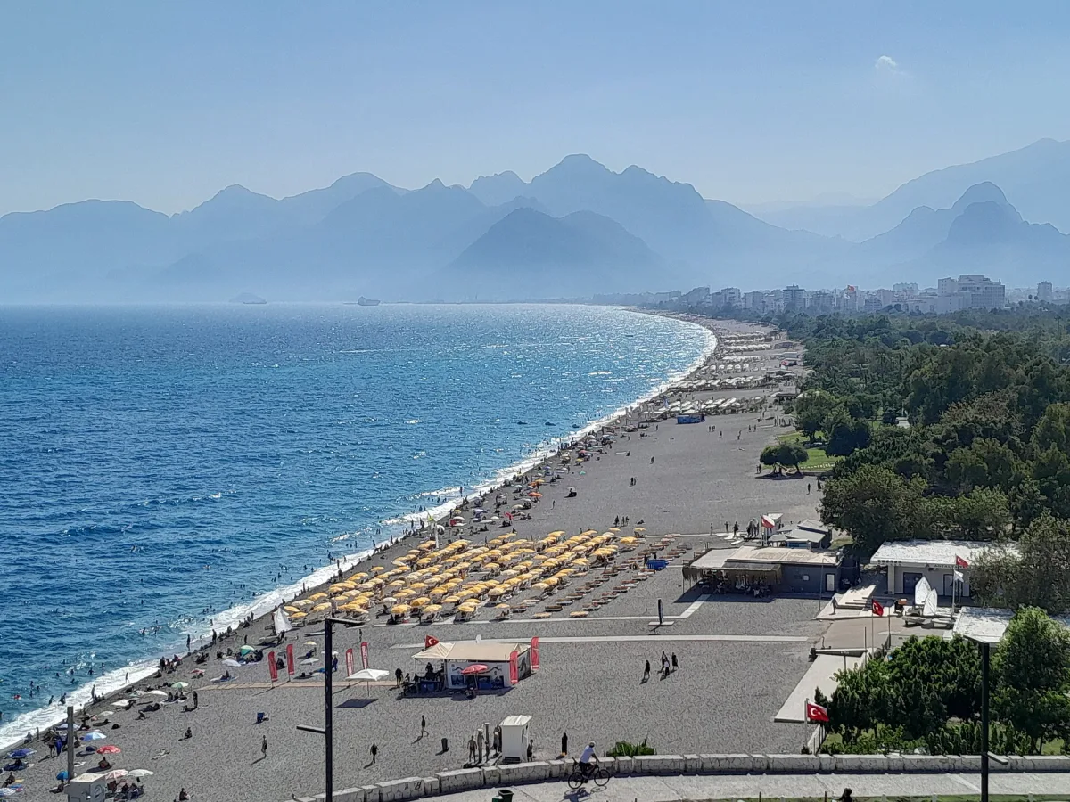 Longue plage rectiligne au revêtement gris bordée de parasols jaunes, mer bleue et chaîne de montagnes dans la brume