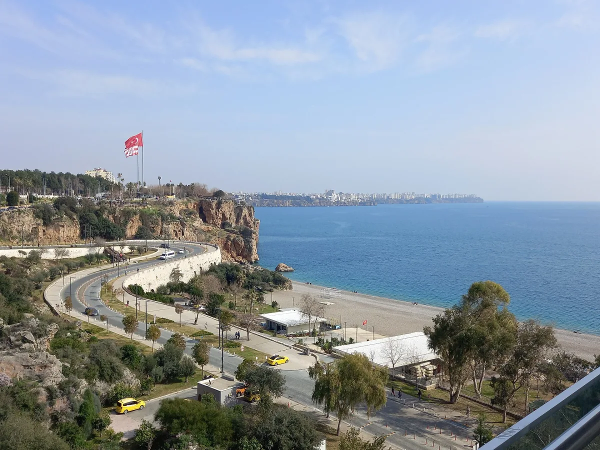 Falaises calcaires surmontées d'un grand drapeau turc, route en lacets et plage de galets pâles en contrebas