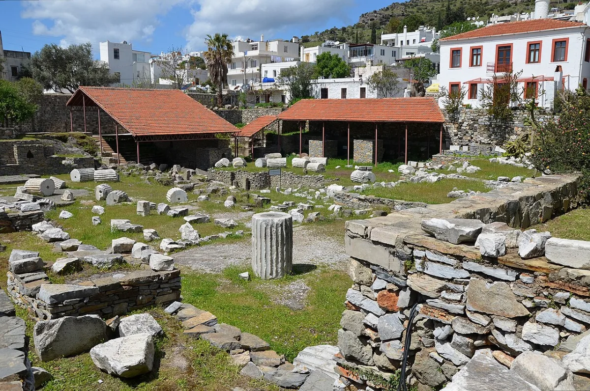 Terrain de fouilles avec fondations, tambours de colonnes et blocs de marbre, maisons blanches en arriere-plan