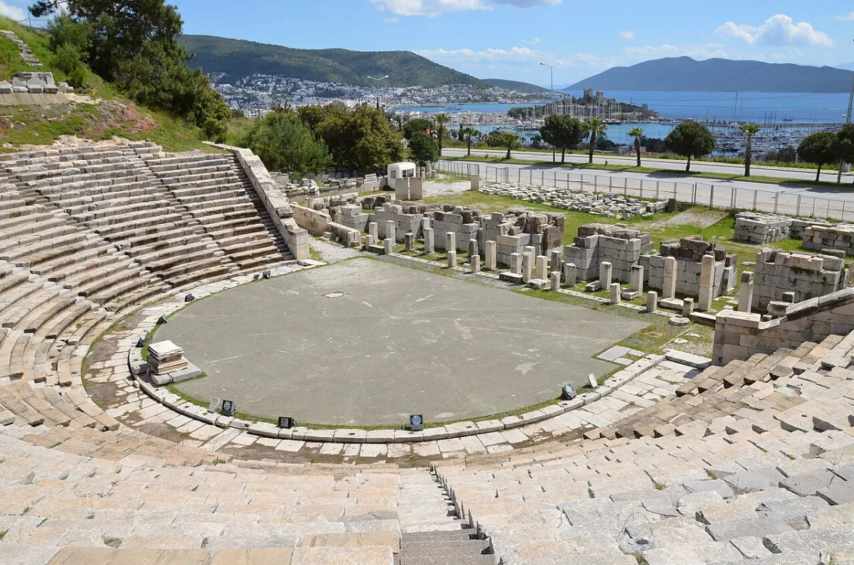 Gradins en demi-cercle d'un théâtre antique dominant une baie et une ville blanche