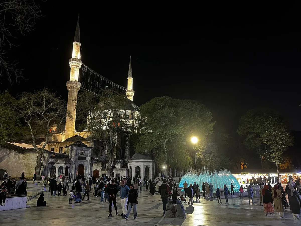 Place bondée devant la mosquée d'Eyüp Sultan à Istanbul le soir, minarets éclairés et fontaine illuminée